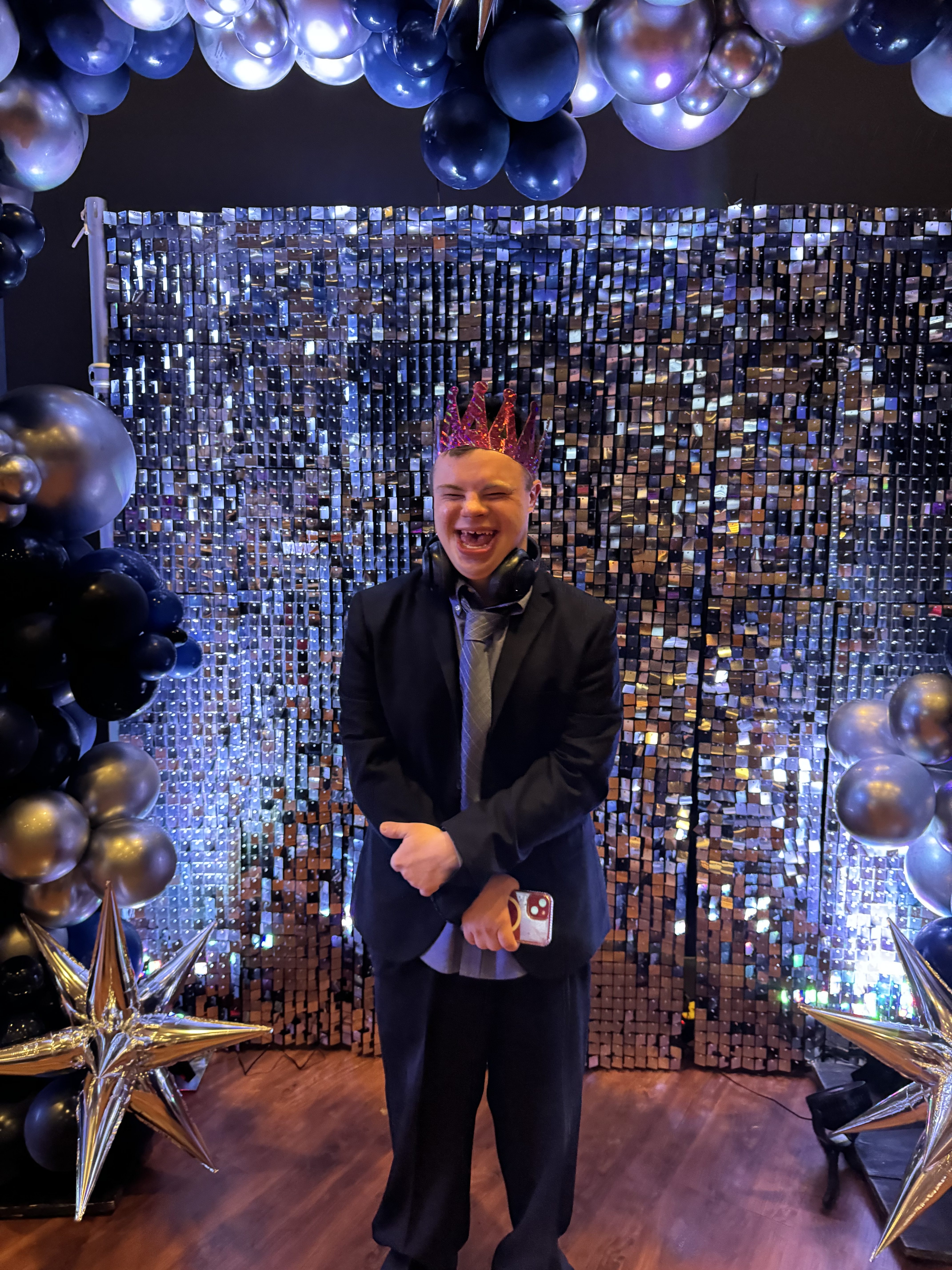 A man in a black suit smiles in front of a shimmering backdrop at a party.