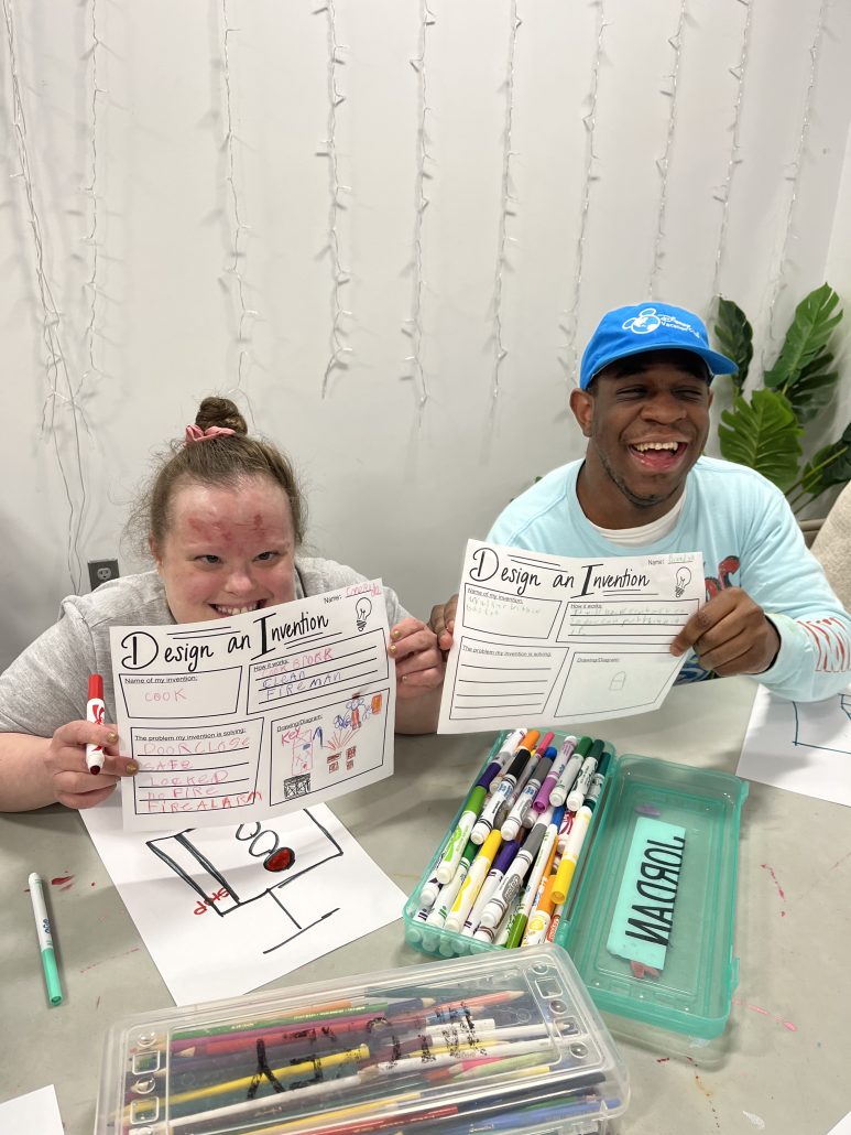 Two people smile while holding up paper that reads "design an invention". They are sitting at a table with containers full of washable markers.