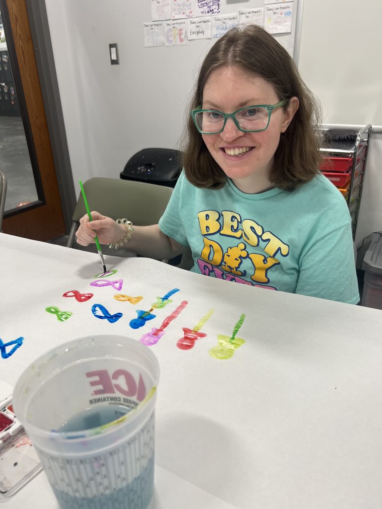 A woman in green glasses smiles and paints on a table.