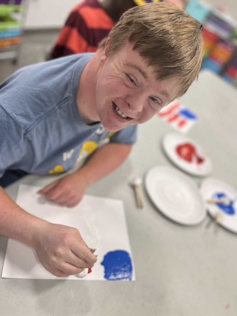 A boy in a blue shirt smiles and paints a piece of paper.